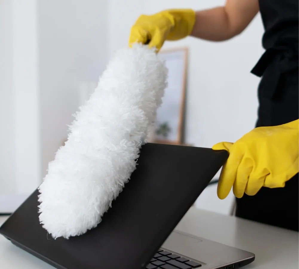 A woman wearing yellow rubber gloves using a white duster to clean a black laptop.