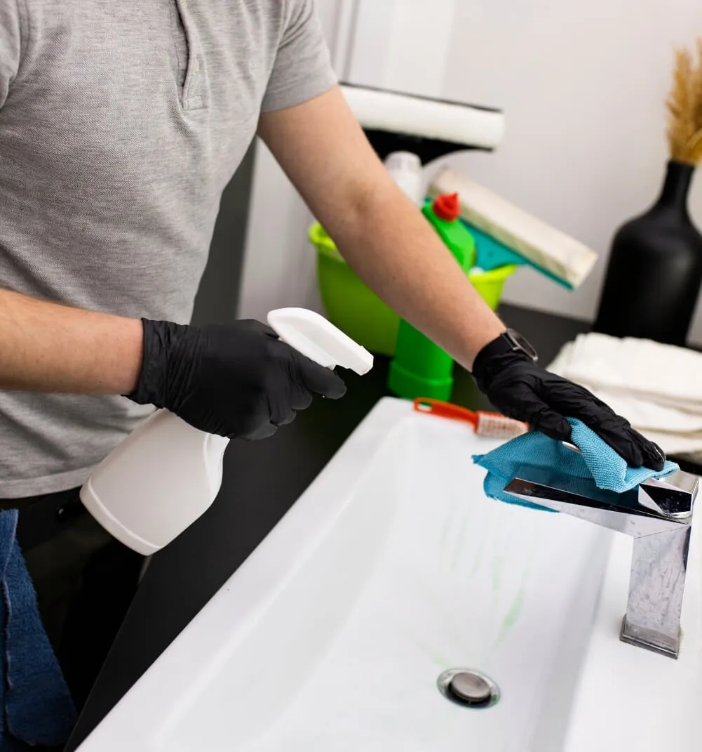 A man wearing black nitrile gloves using a white water bottle and blue microfiber cloth to clean a stainless steel faucet.