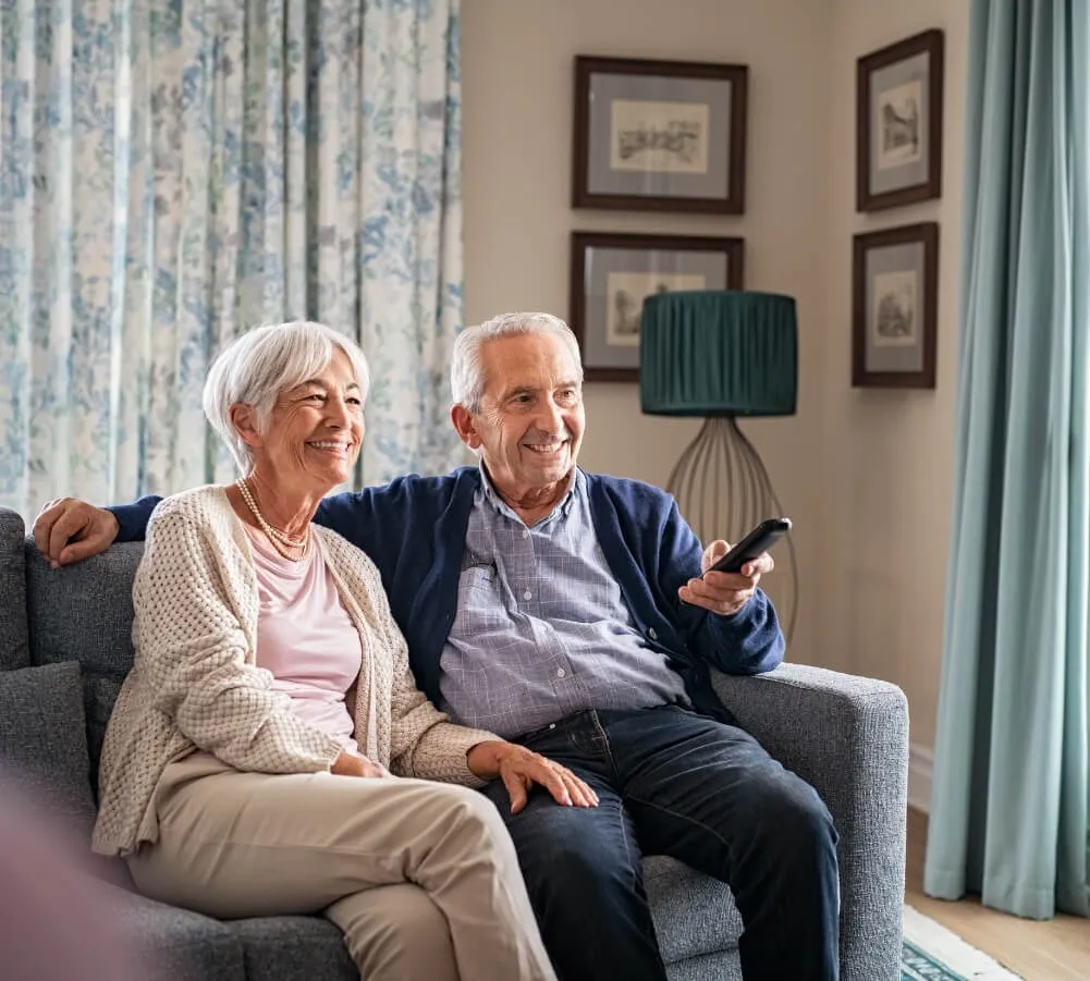 An elderly man and wife sitting together on a sofa are smiling and enjoying a clean home that was cleaned by Sparkle n' Clean.