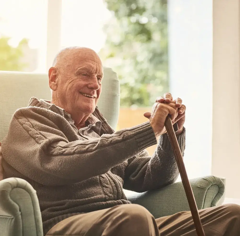 An elderly man with a cane sits on a green chair and smiles as he takes in the sight of a room cleaned by Sparkle n' Clean.